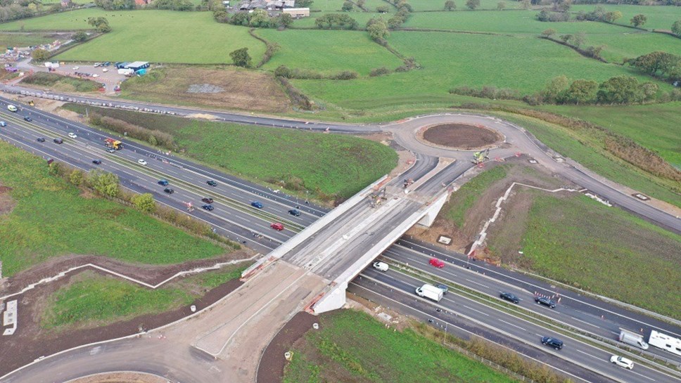 Aerial view of a new road bridge and roundabout under construction beside a busy motorway in open countryside